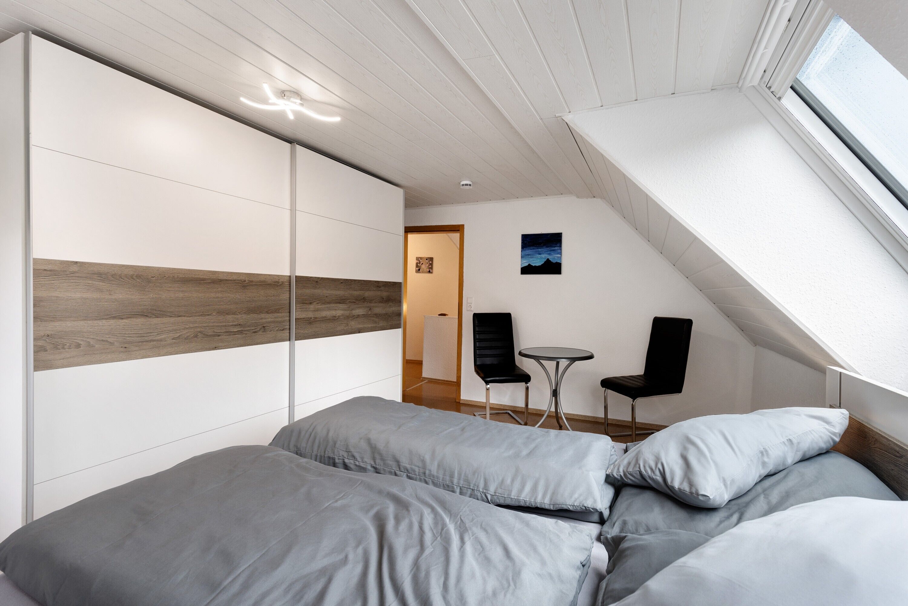 Ein Schlafzimmer mit schrägen Wänden, einem Bett mit grauer Bettwäsche, einem kleinen Tisch, zwei Stühlen und einem Bild an der Wand.