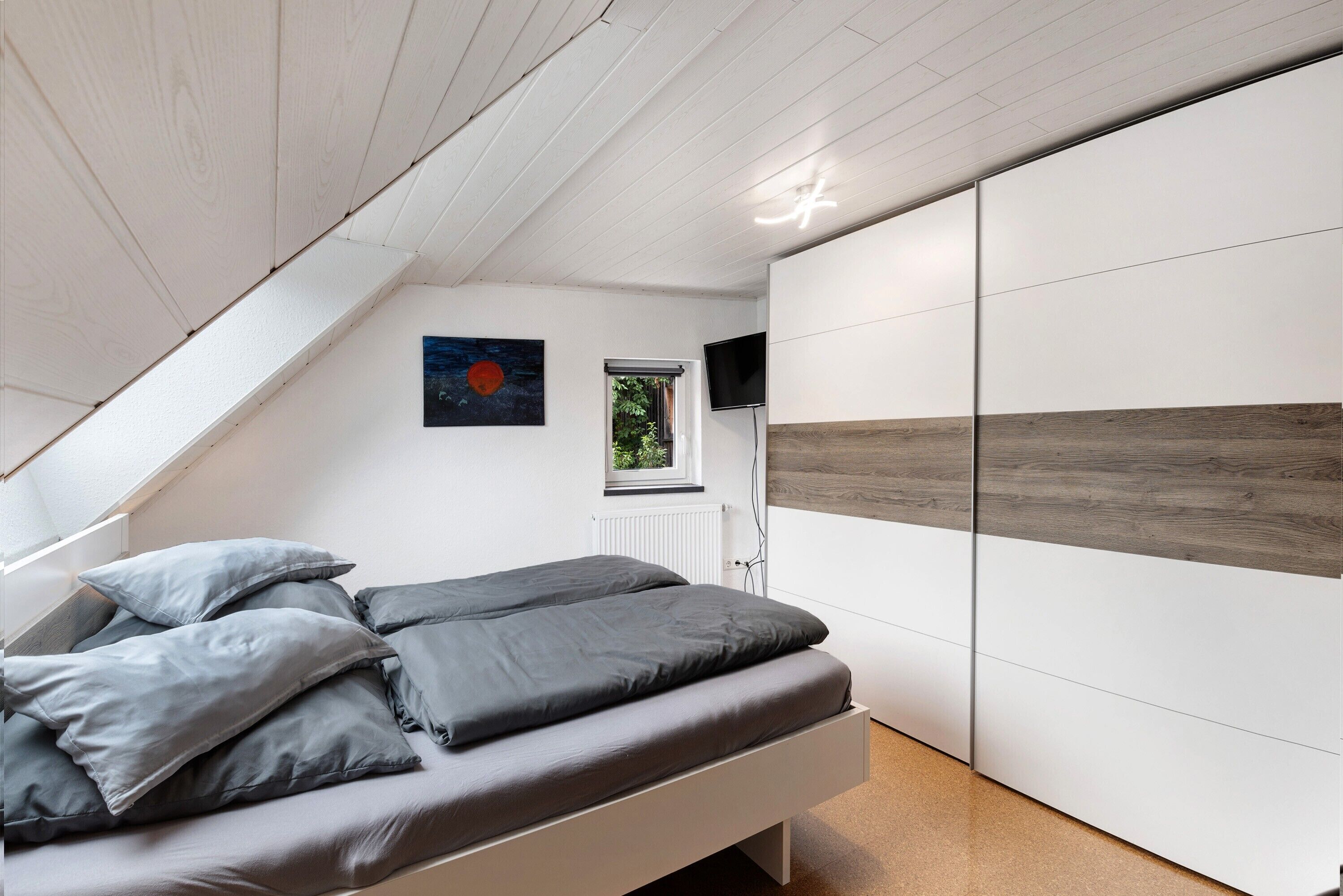 Quarto com teto inclinado, cama com roupa de cama cinza, televisão fixada na parede e janela com vista para o verde.