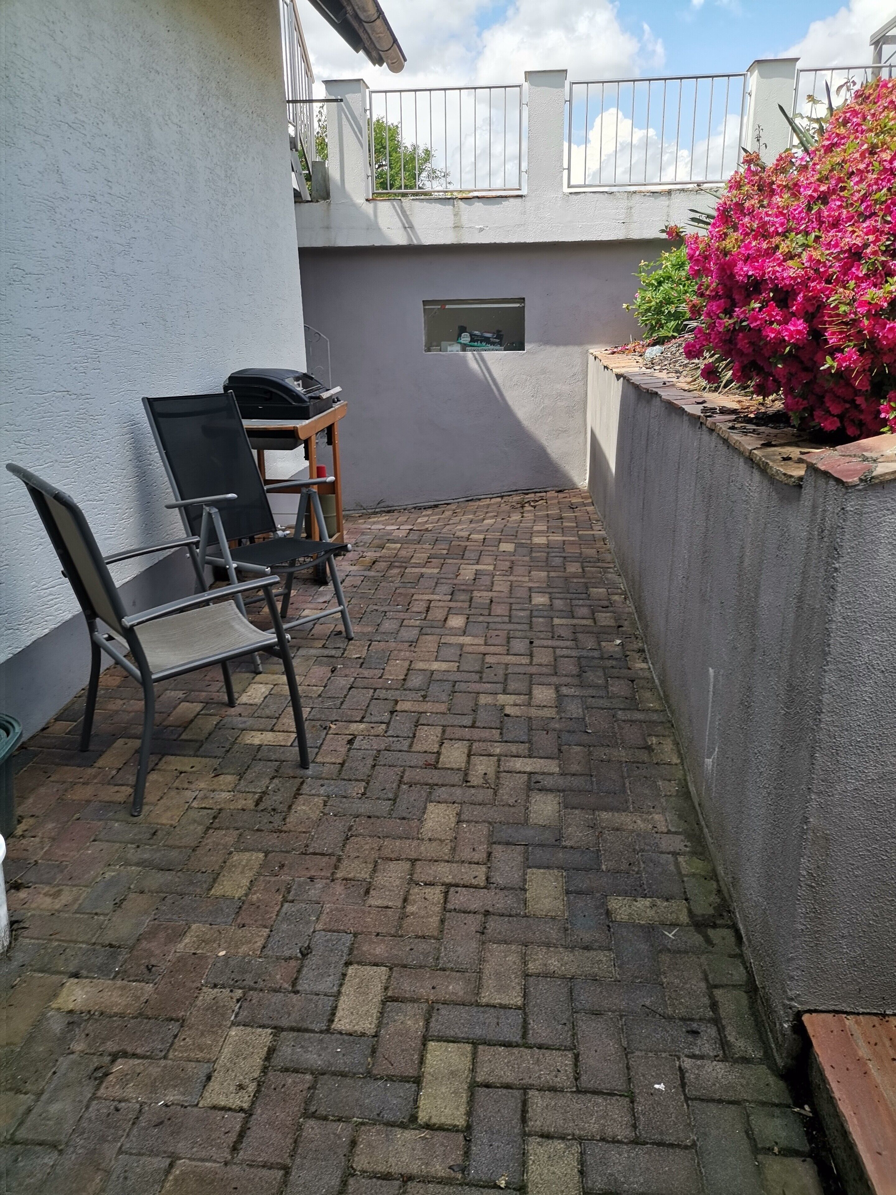 Un'area patio con pavimento in mattoni, una sedia in metallo, un tavolino e un aiuola con fiori rosa.