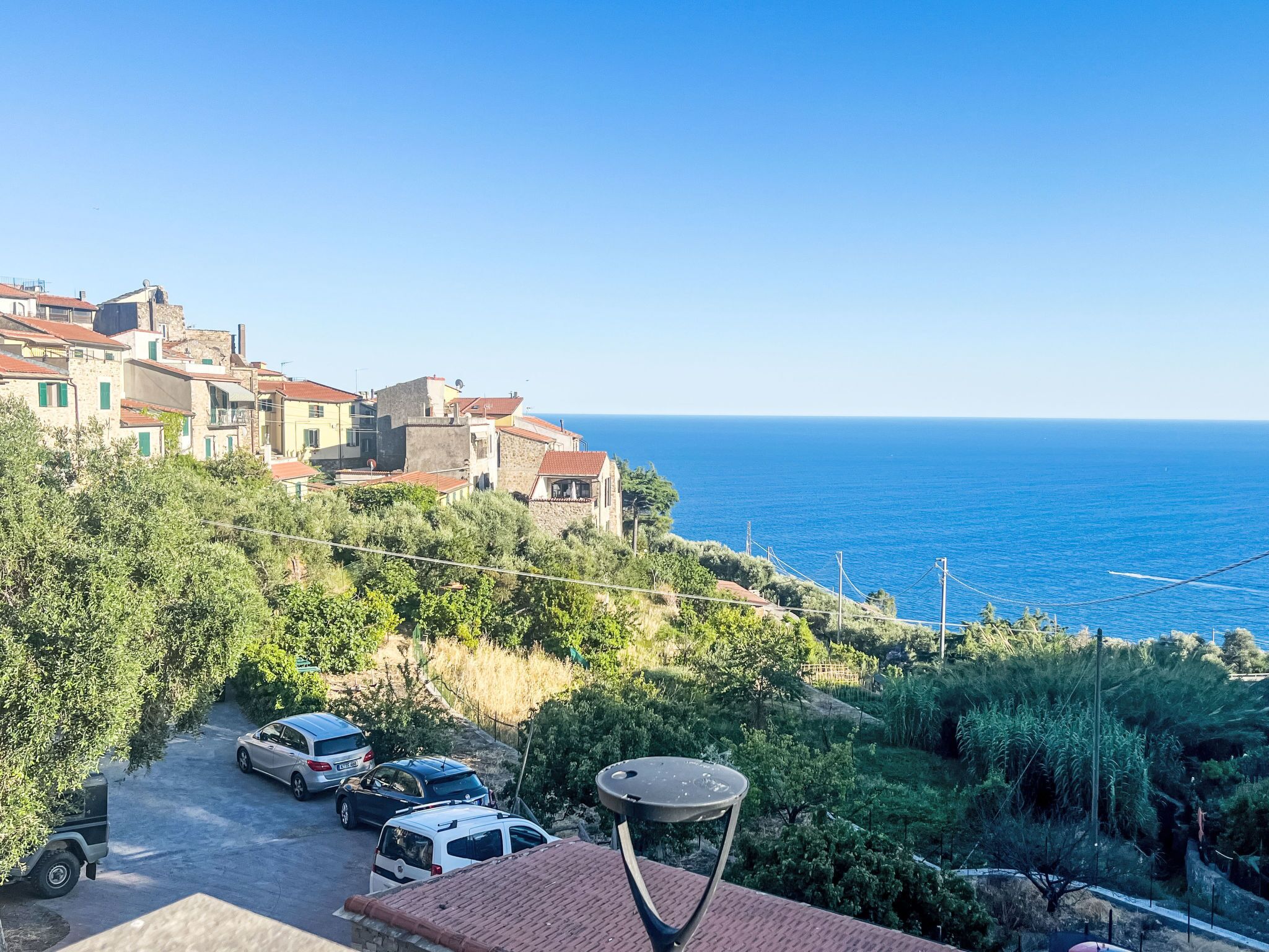 A coastal village with traditional stone buildings, a paved road, parked cars, and a clear view of the sea.