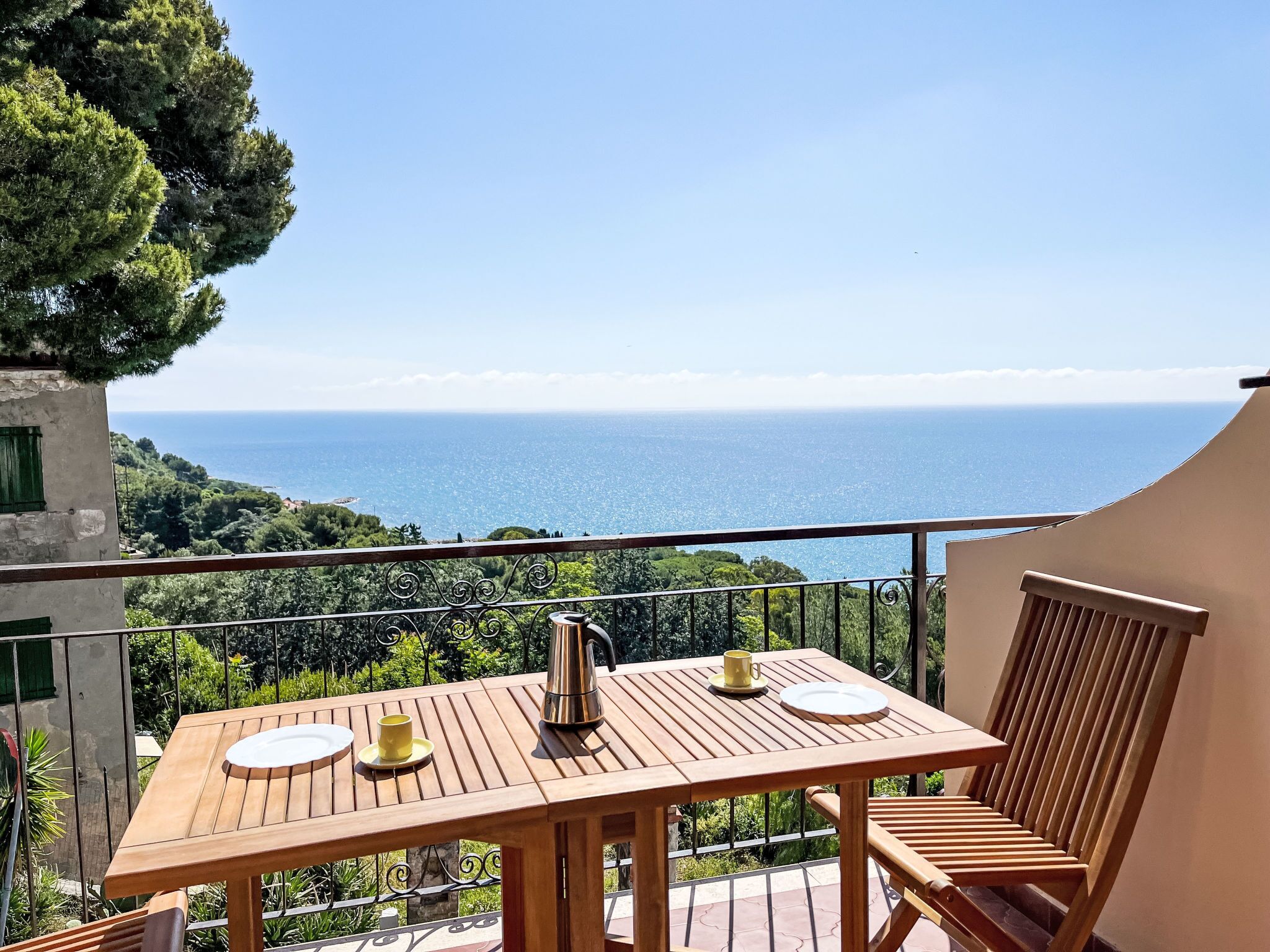 Une table en bois, une bouilloire en métal et une tasse jaune sont posés sur un balcon donnant sur la mer.