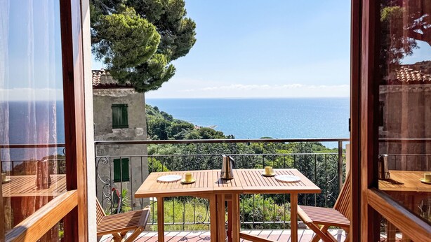 A balcony with a wooden table and chairs, overlooking a coastal landscape with a view of the sea and greenery.