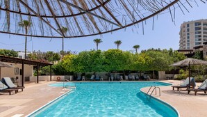A swimming pool with lounge chairs and umbrellas, surrounded by greenery and a building in the background. - Phaedrus Living : Limassol Pyrgos Seaside Flat (Pyrgos)