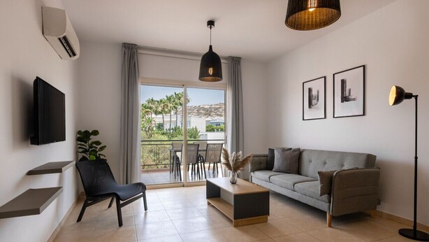 A modern living room with a grey sofa, a black chair, a wooden coffee table, and a floor lamp. There is a TV mounted on the wall, a plant on a shelf, and a balcony with a view of palm trees and buildings. - Phaedrus Living : Limassol Pyrgos Seaside Flat (Pyrgos)