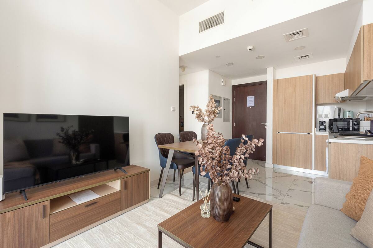 Ein modernes Wohnzimmer mit Flachbildfernseher, Holzmöbeln, einer Vase mit Trockenblumen und einer Küchenzeile mit Holzausstattung und Geräten.