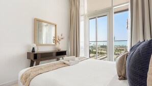 Ein Hotelzimmer mit einem großen Bett, einem Nachttisch mit Vase und Lampe, einem Spiegel und Blick auf die Stadt durch große Fenster.