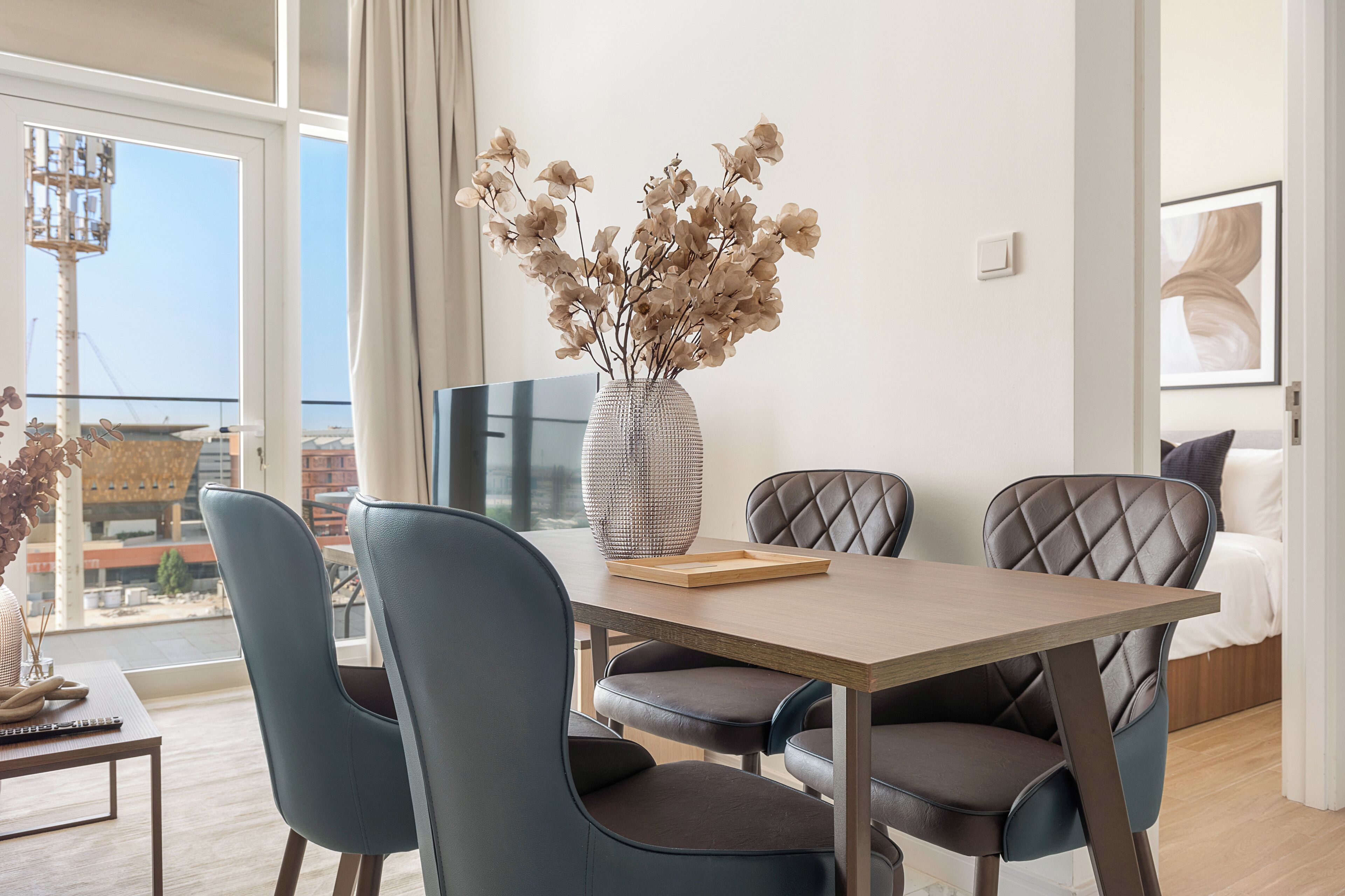 A modern dining area with a wooden table, cushioned chairs, and a vase with dried flowers.