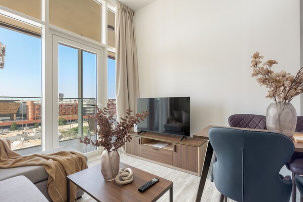 Ein modernes Wohnzimmer mit großem Fenster, einer Couch, einem Couchtisch, einem Fernseher und einer Vase mit Trockenblumen.