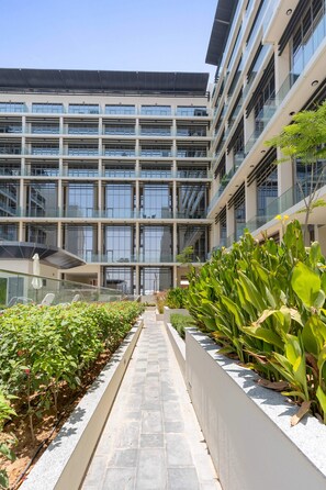 Un edificio moderno de varios pisos, con amplias ventanas y un camino central bordeado por jardineras con follaje.