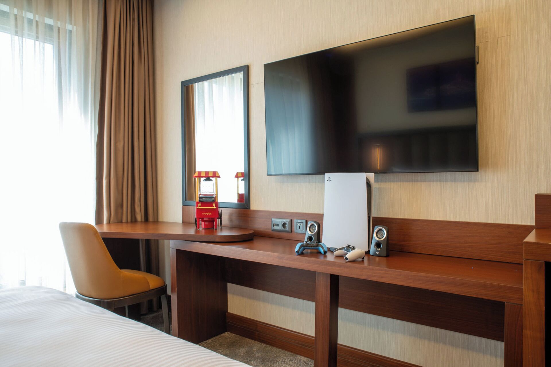 Habitación de hotel con un escritorio de madera, un televisor montado en la pared, una cama con edredón blanco y una ventana con cortinas.