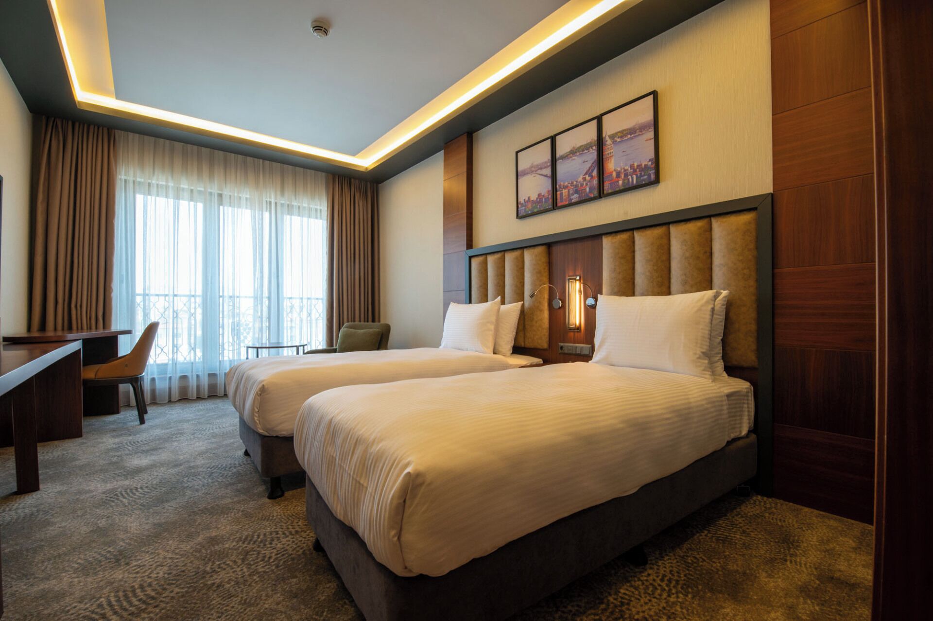 A hotel room with two beds, a desk, a chair, and a large window with curtains.