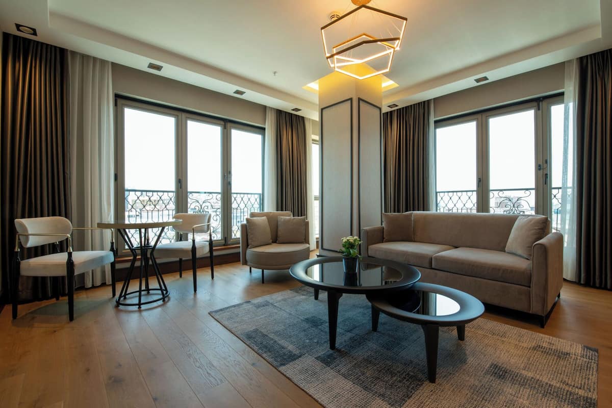 A modern living room with a sofa, two armchairs, a coffee table, and a dining table. Large windows offer a view of the city.