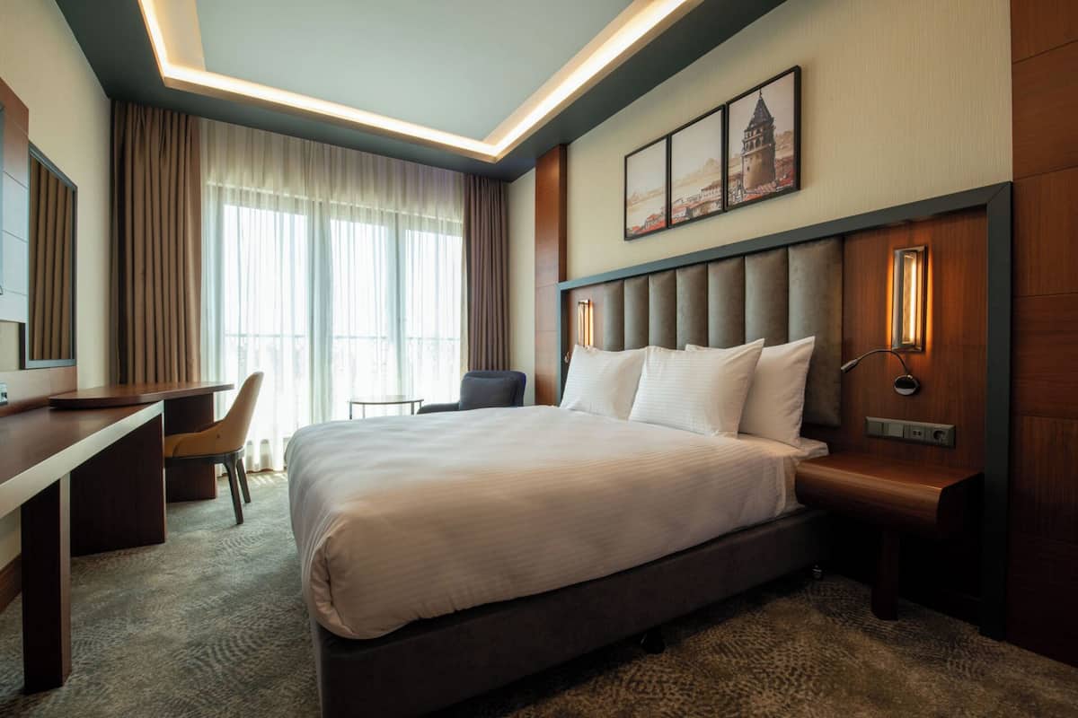 A hotel room with a large bed, a desk, a chair, and a view of the city through the window.