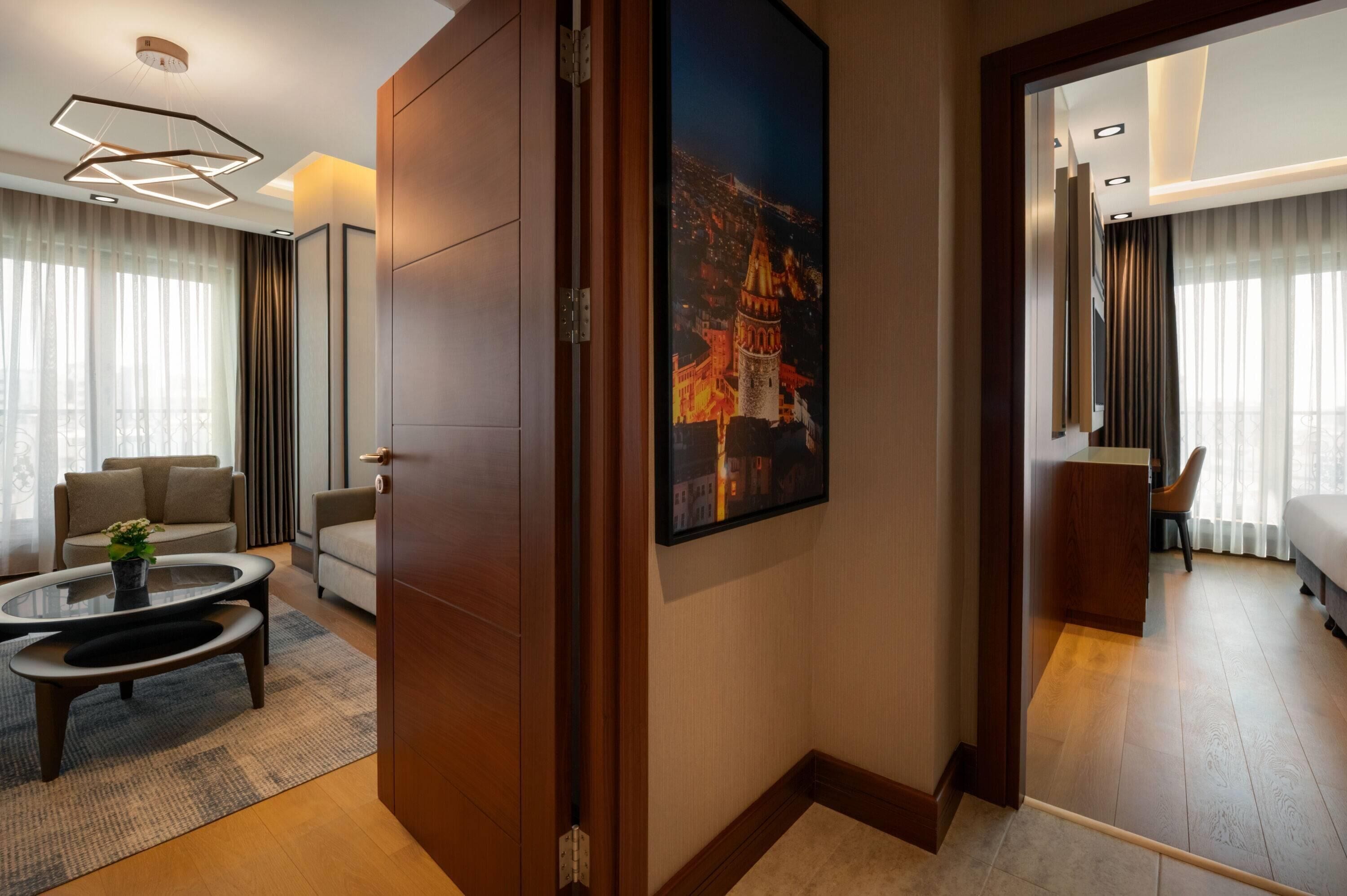 A modern hotel room with a large bed, a sitting area with a sofa and a coffee table, a painting of a cityscape, and a wooden door.