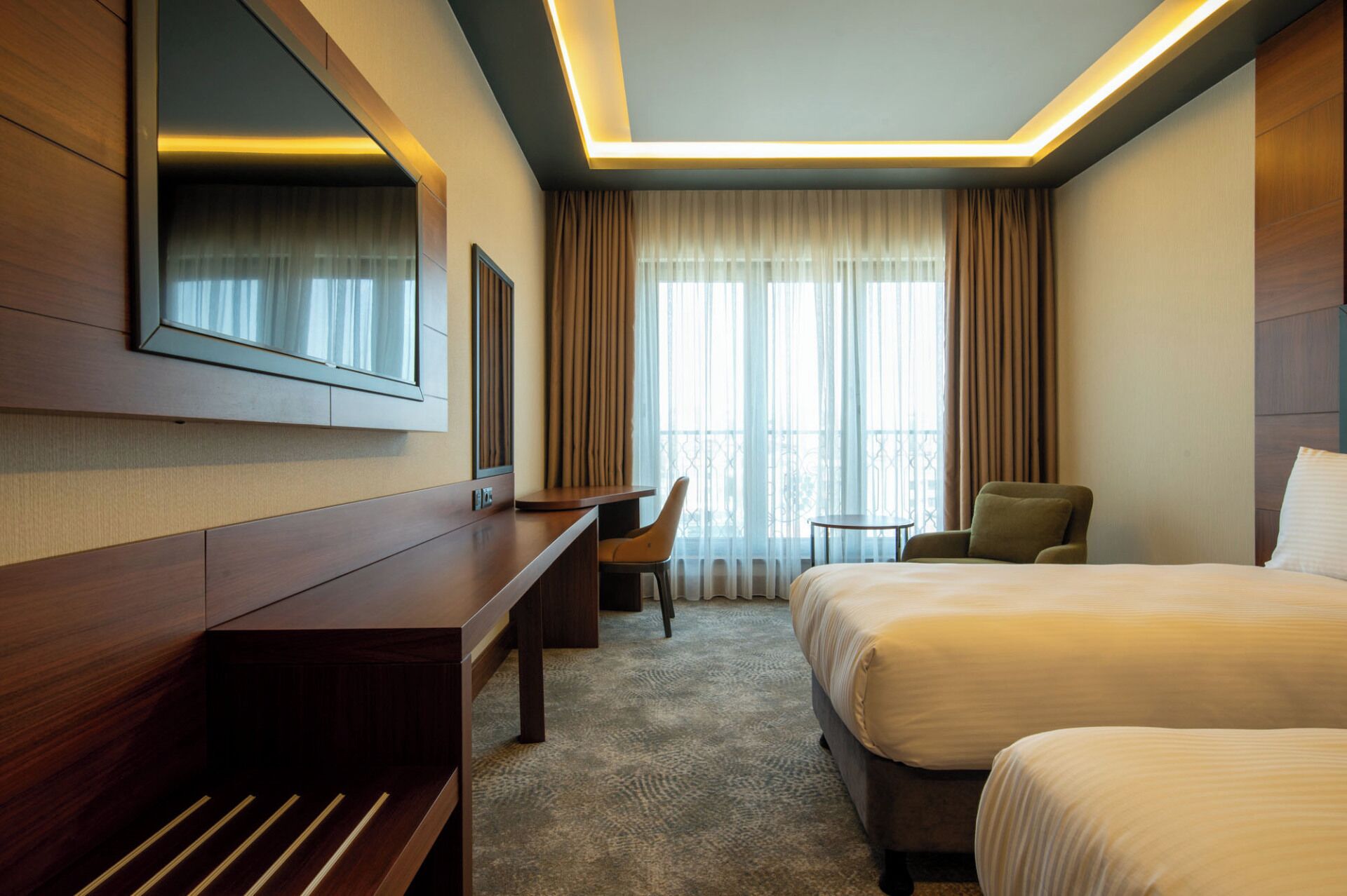 A hotel room with a bed, a desk, a chair, a small table, a TV, and a large window with curtains.
