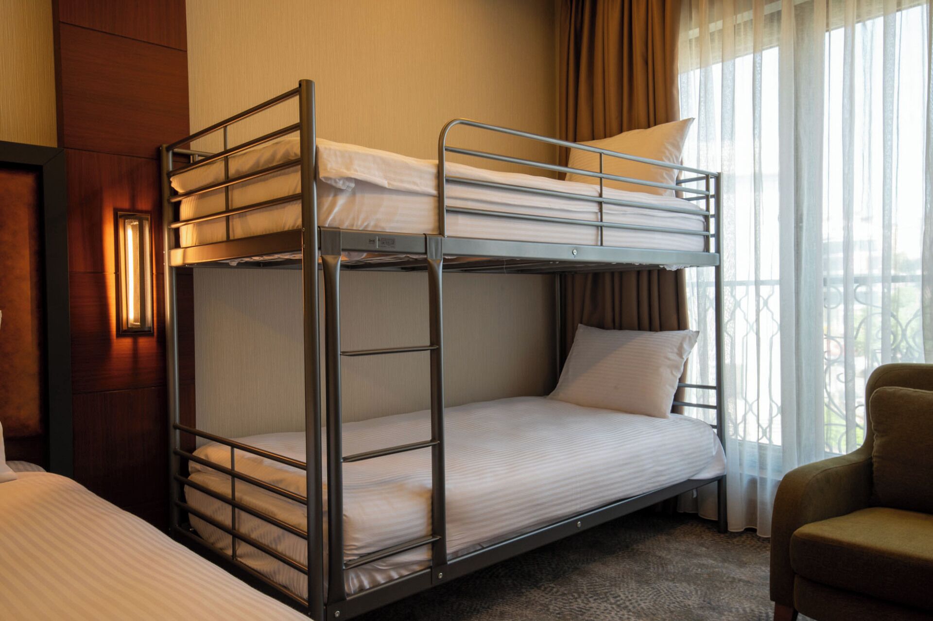 A bunk bed in a hotel room with a window, curtains, and a chair.