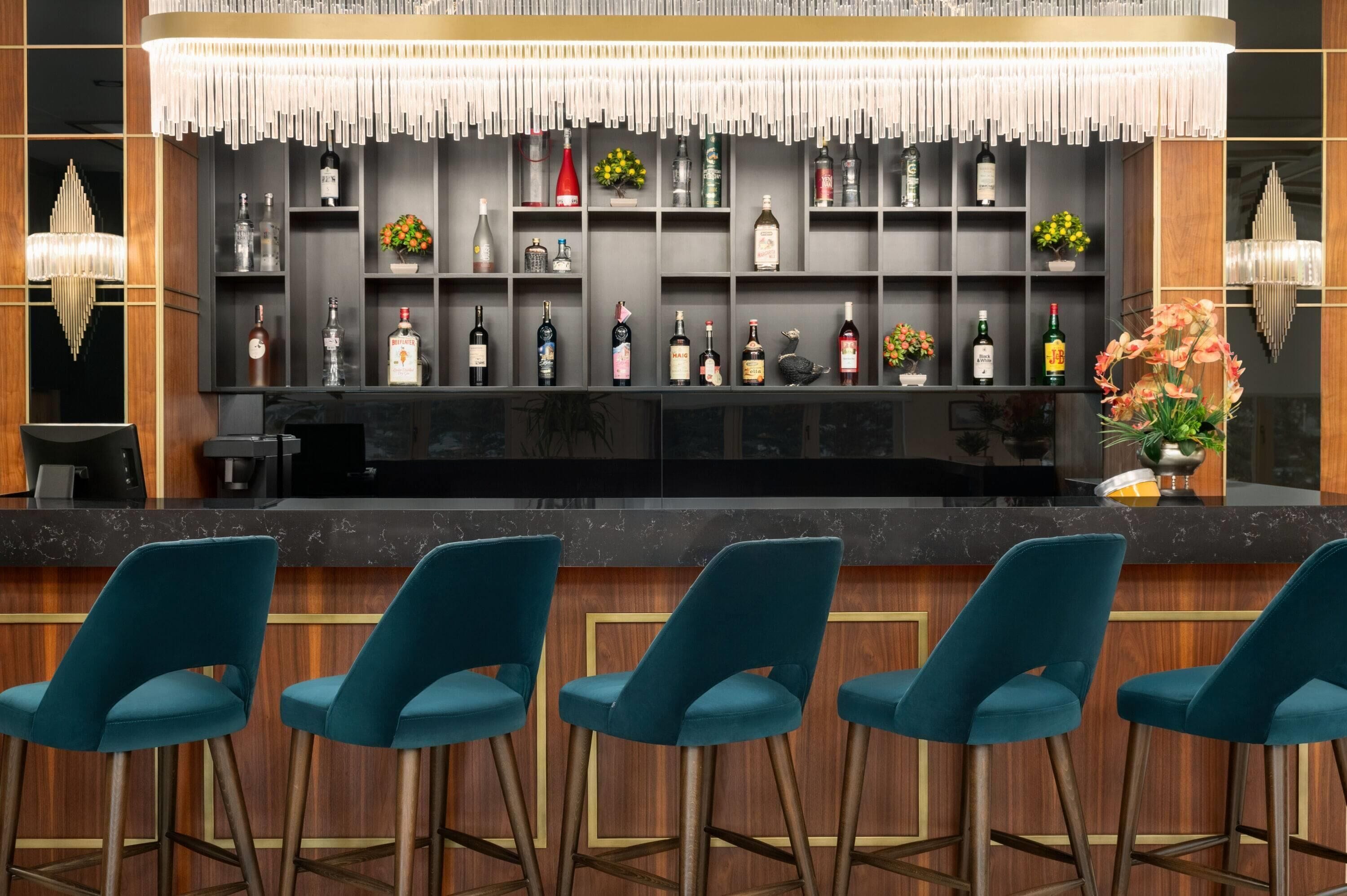 Uma área de bar com bancada de mármore, banquetas azuis-turquesa e diversas garrafas de bebidas alcoólicas nas prateleiras.