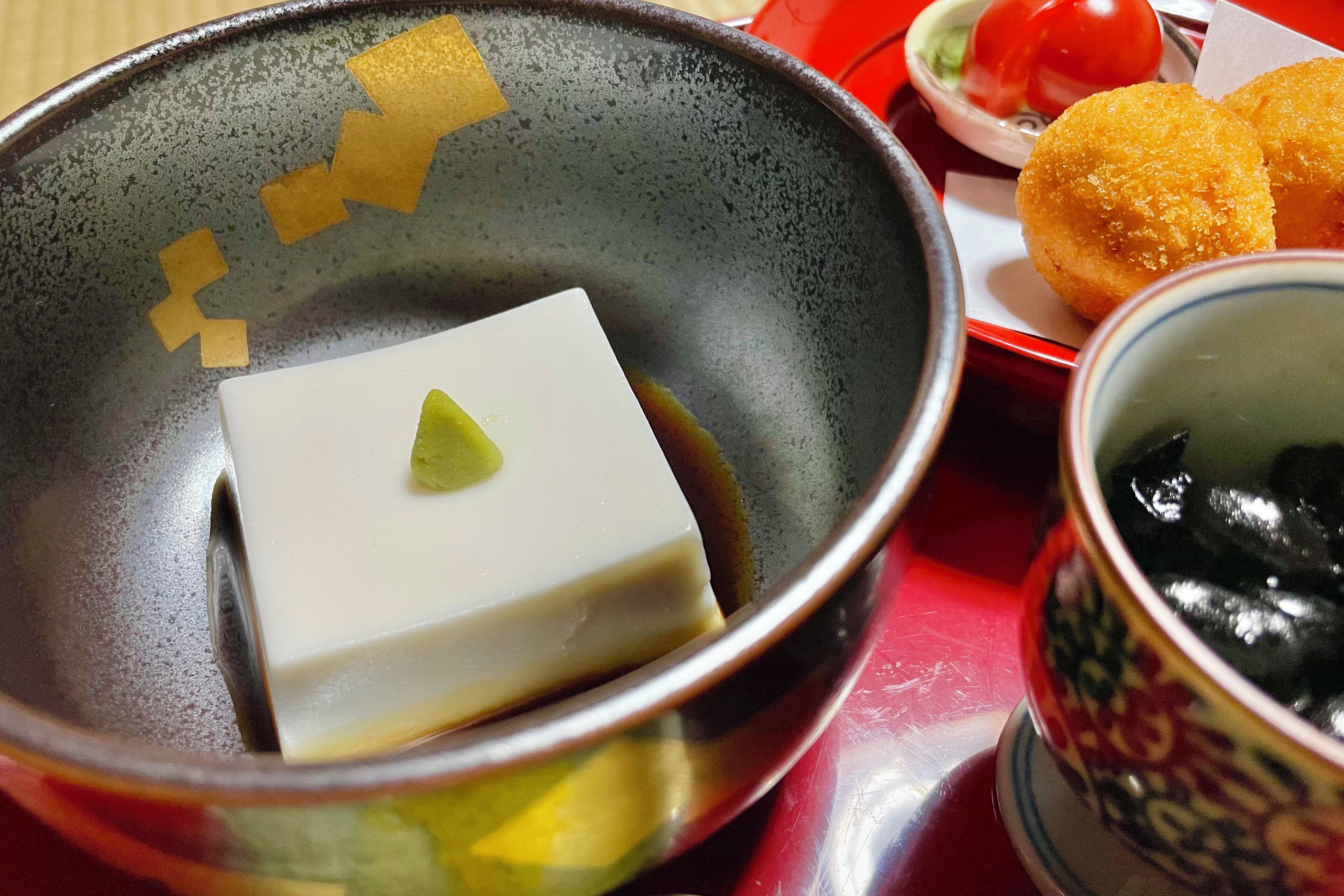 Un tazón con un bloque de tofu y un vegetal verde pequeño, una taza con contenido negro, y un plato con dos alimentos fritos.