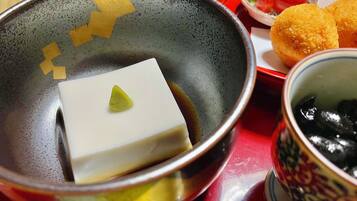 Un tazón con un bloque de tofu y un vegetal verde pequeño, una taza con contenido negro, y un plato con dos alimentos fritos.