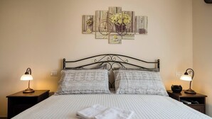 A neatly made bed with a white quilt, two pillows, and a folded towel. There are two bedside lamps on wooden nightstands. A multi-panel artwork is above the bed. - Villa Clara Alberobello (Alberobello)