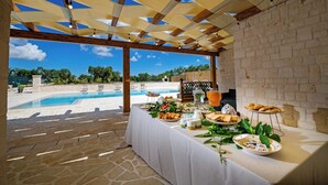 A covered outdoor dining area with a pool view, stone flooring, and a table set with breakfast items. - Villa Clara Alberobello (Alberobello)
