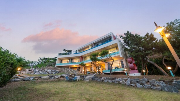 A modern multi-story building with a flat roof, outdoor seating, and a stone retaining wall. - Ganghwado Happyvilly Pension (Incheon)