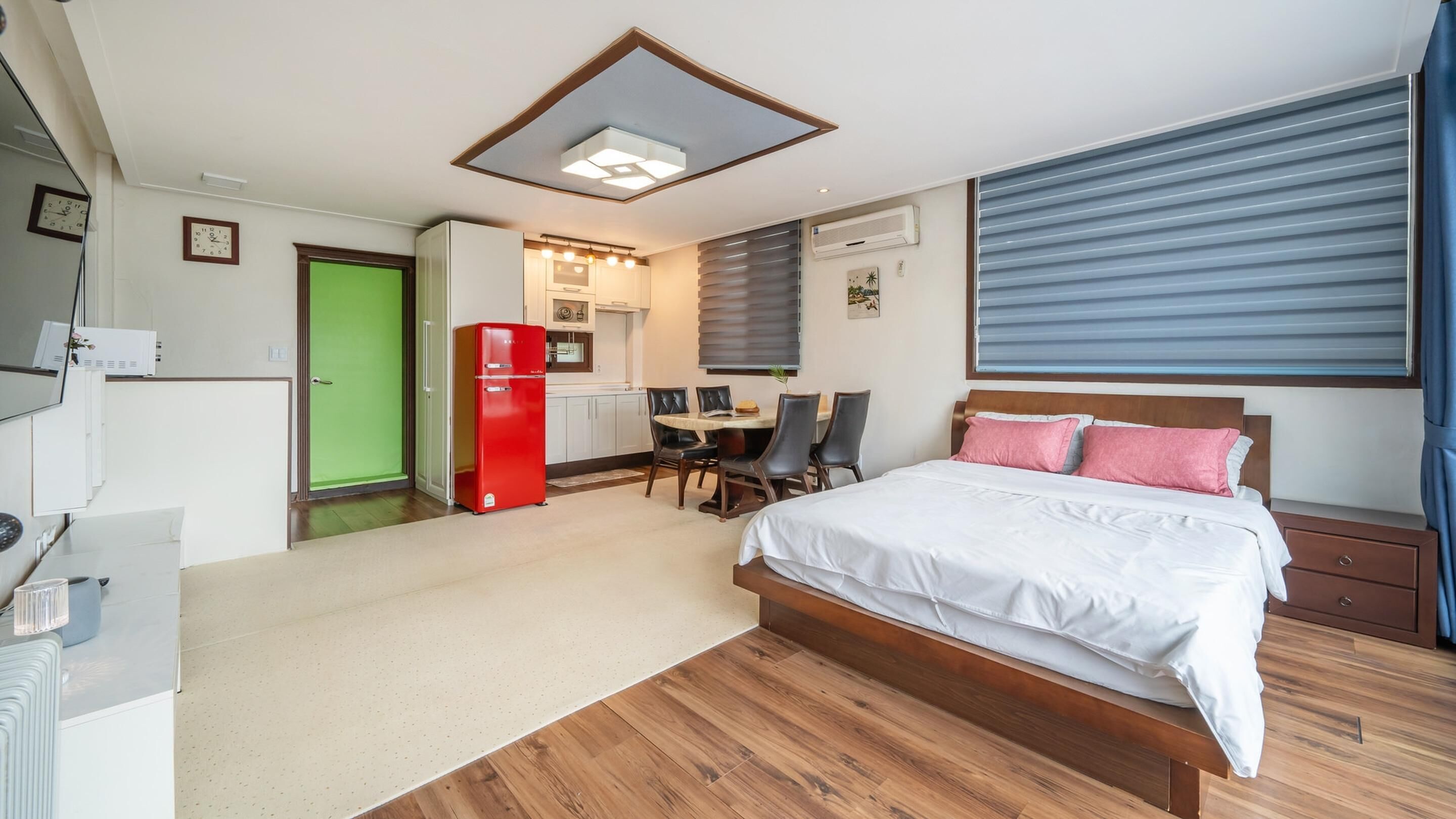 A hotel room with a bed, a desk, a red refrigerator, a dining area, and a green door.