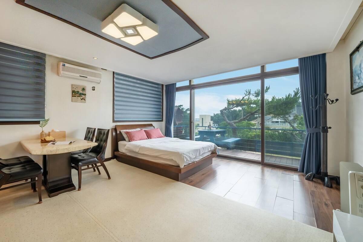 Une chambre d’hôtel moderne, équipée d’un lit, d’un bureau et d’une chaise. Une grande fenêtre offre une vue sur l’extérieur.