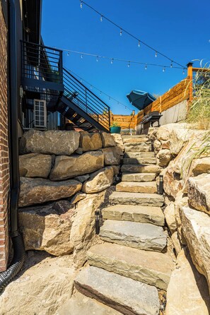 Una escalera de piedra que sube a una terraza de madera con luces colgantes.