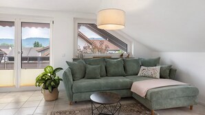 Een groene bank met een witte plaid, een klein rond tafeltje en een potplant in een kamer met uitzicht op huizen en bergen door een raam.