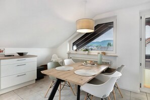 Une cuisine moderne avec une table en bois, des chaises blanches, un luminaire suspendu et une fenêtre donnant sur les montagnes.