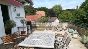 Eine Holzterrasse mit Tisch und Stühlen, Blumentöpfen und Blick auf ein Haus im Hintergrund.