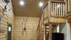 A log cabin interior with a wooden staircase, a sofa with decorative pillows, and a black refrigerator. - Riverfront, Authentic, Luxury Log Cabin-Bison (Wasilla)