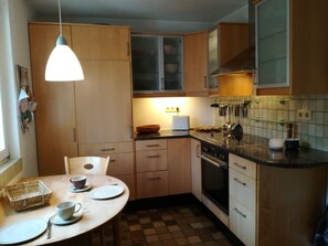 A compact kitchen with wooden cabinets, a dining table set for two, a built-in oven, and a hanging light fixture.