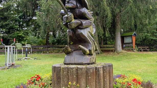 A sculpture of a figure with a staff, placed on a wooden stump with flowers around it.