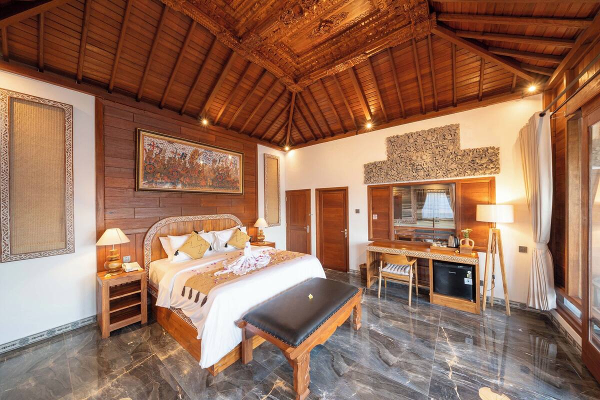A spacious bedroom with a large bed, a desk, and a chair, wooden ceiling, and marble flooring.