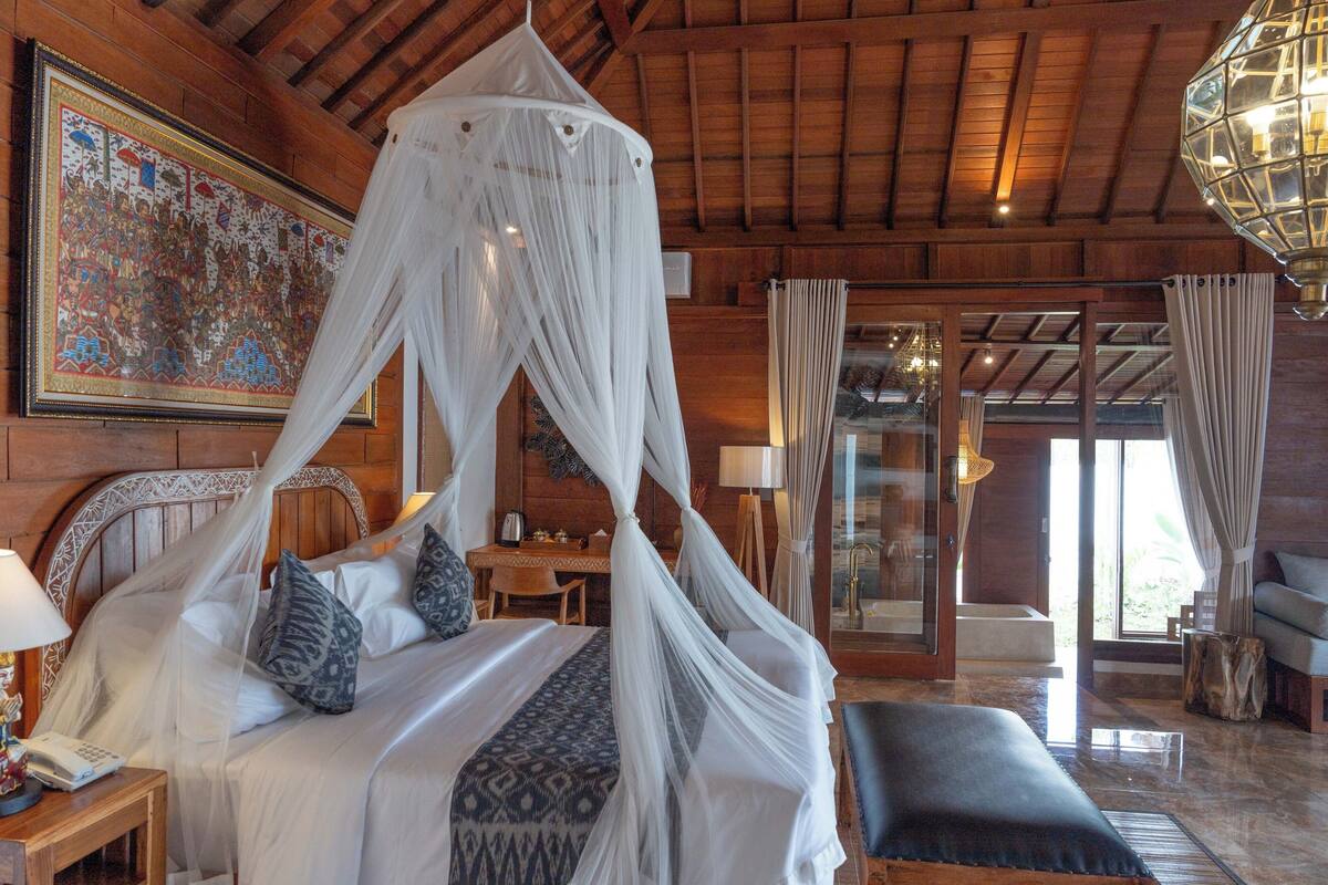 A room with a bed under a canopy, a wooden ceiling, a large framed artwork, and a view through a glass door.