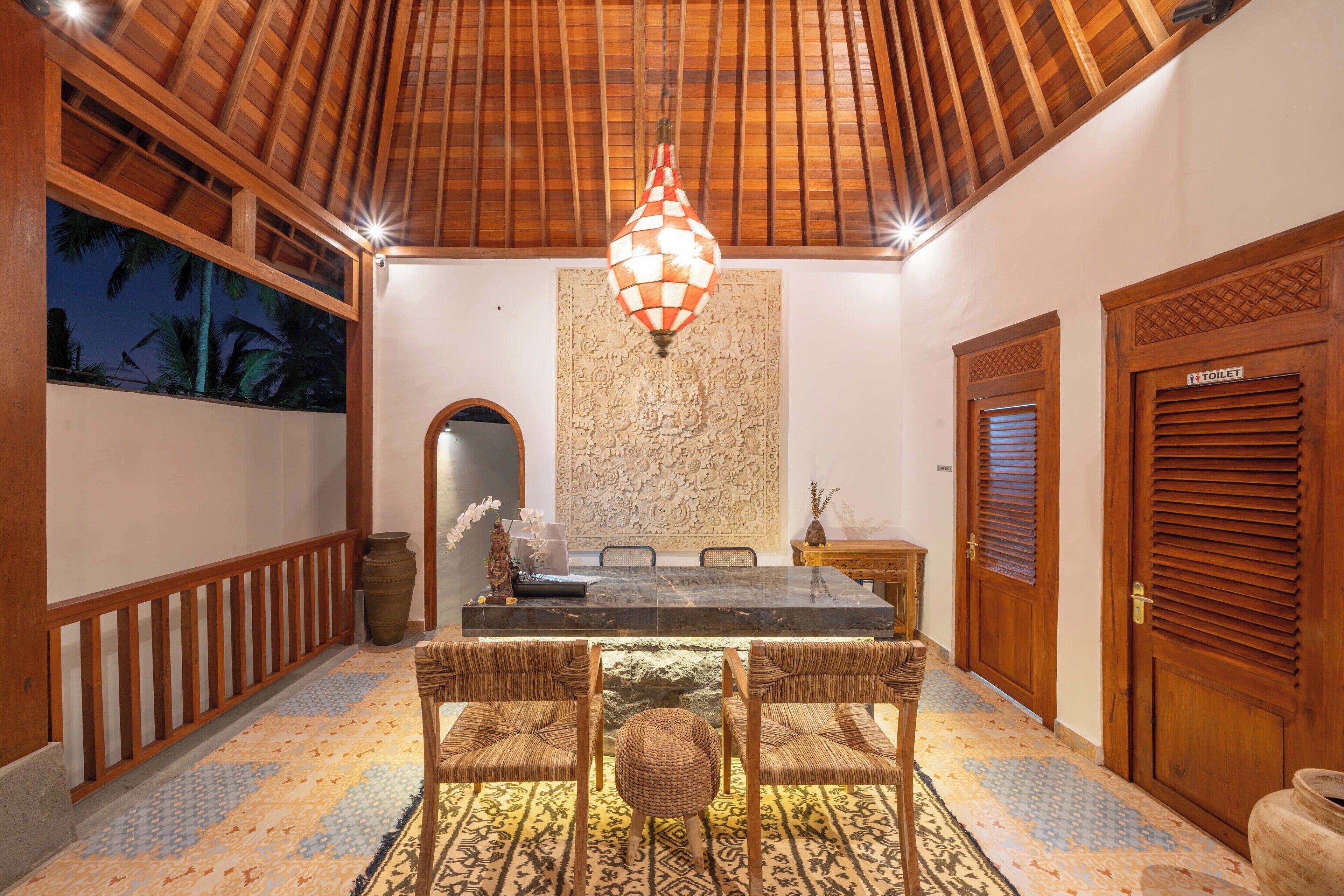 A dining area with a wooden ceiling, a checkered hanging light, a large wall tapestry, and a sign indicating a toilet.
