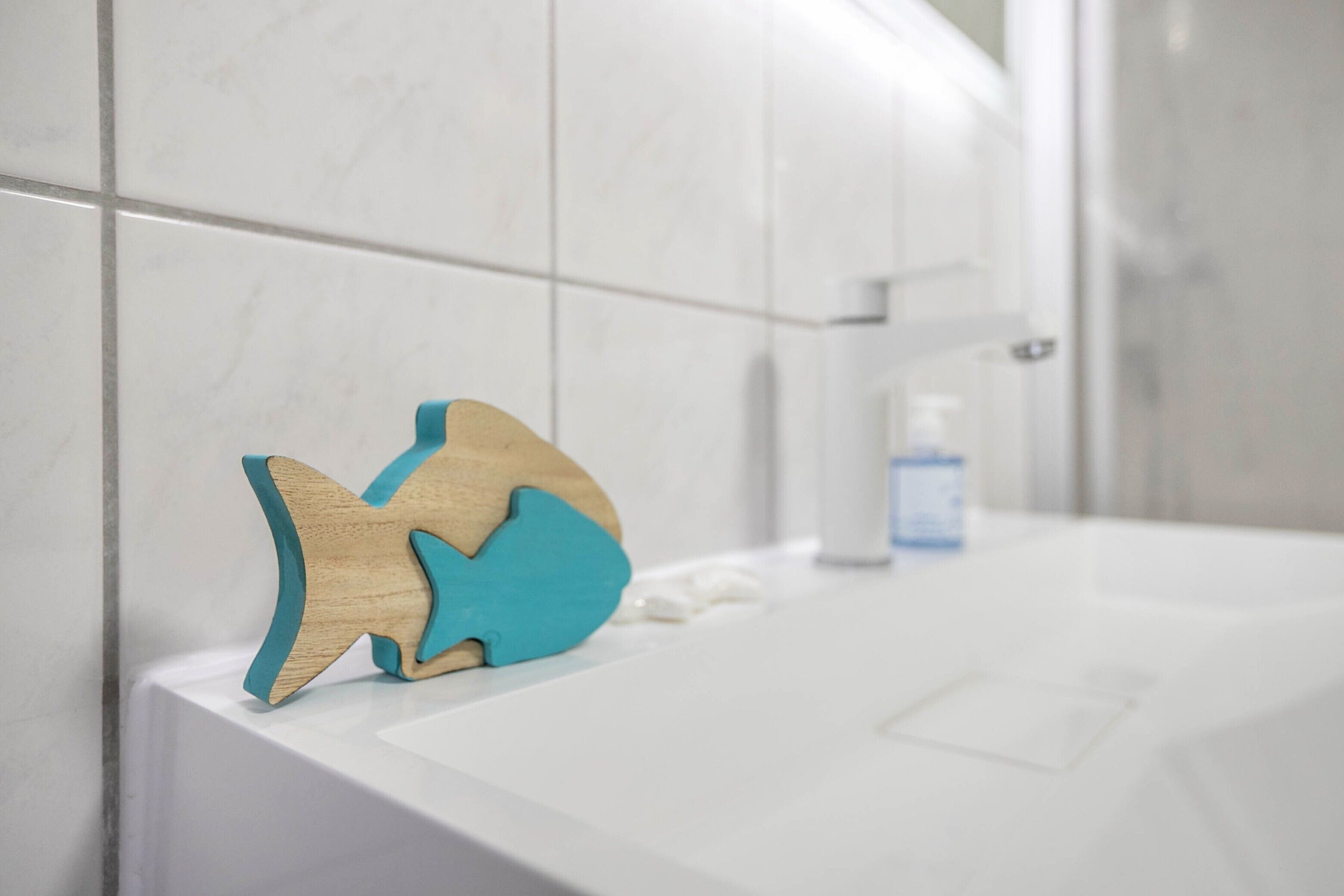 A bathroom with white tiles, a wooden fish-shaped soap dish, and a bottle of soap on the counter.