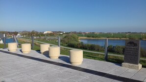 A paved walkway with bollards, a sign with a house illustration, and a view of a river and fields. - Apartment 'Michelsen' with Private Terrace, Private Garden and Wi-Fi (Nordstrand)