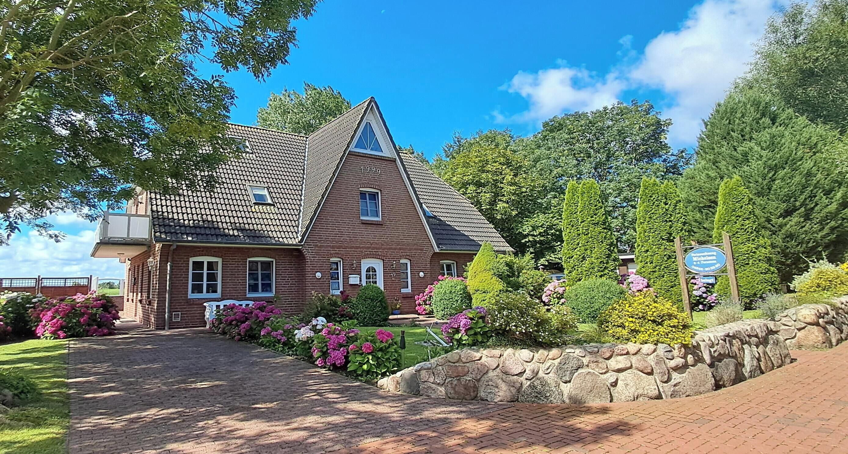 A brick house with a triangular roof, a stone wall, and a garden with various plants and flowers.