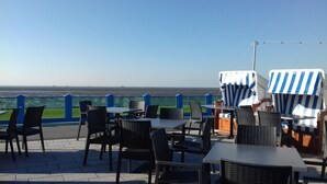 An outdoor seating area with tables and chairs, a view of the sea, and a blue railing. - Apartment 'Michelsen' with Private Terrace, Private Garden and Wi-Fi (Nordstrand)