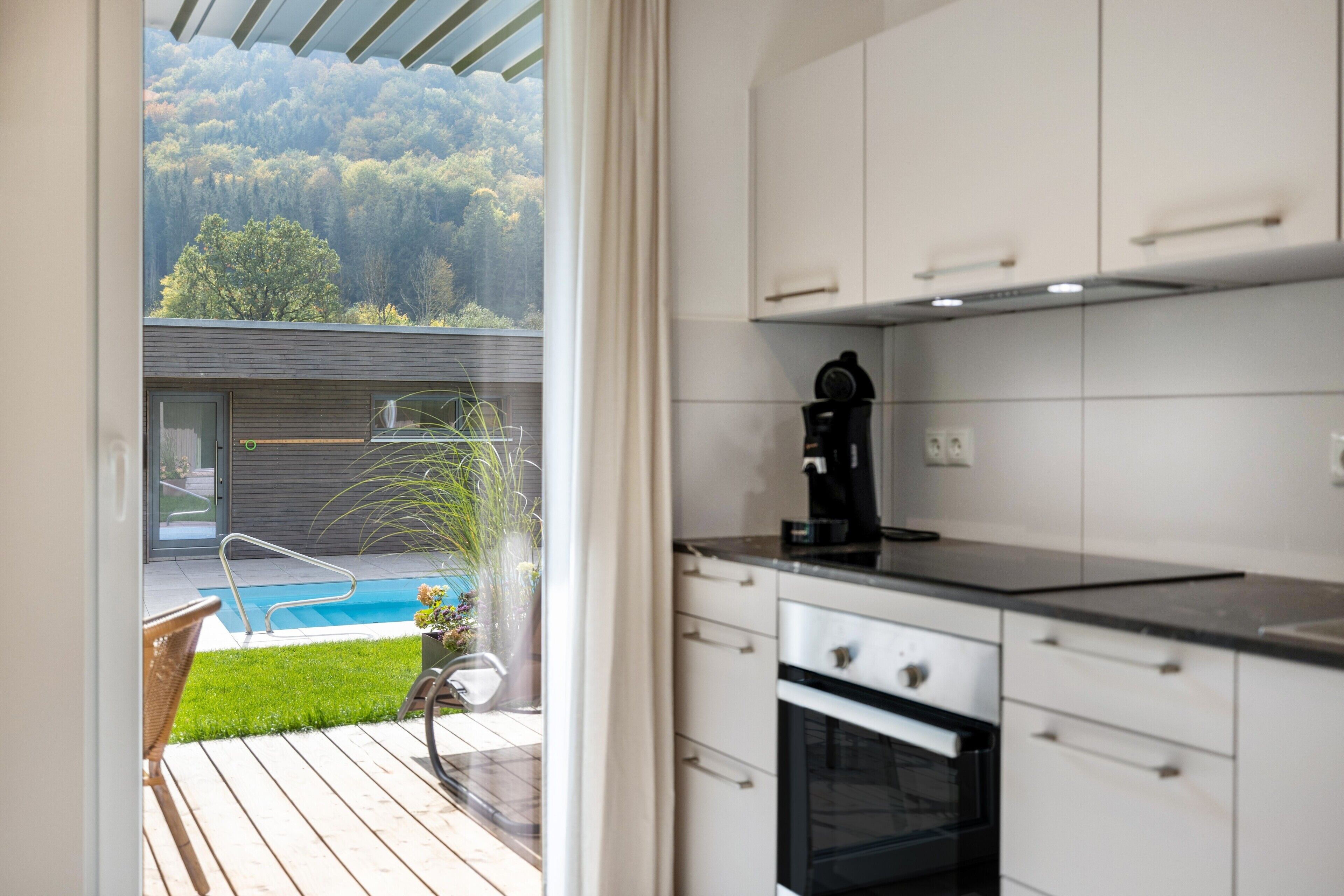 A modern kitchen with white cabinets, a built-in oven, and a view of a swimming pool and garden.