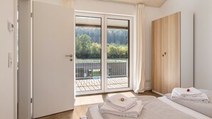 Quarto com cama, guarda-roupa de madeira, porta de vidro deslizante para uma varanda e vista para árvores.