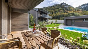 Um deck de madeira com cadeiras de vime, piscina e montanhas ao fundo.