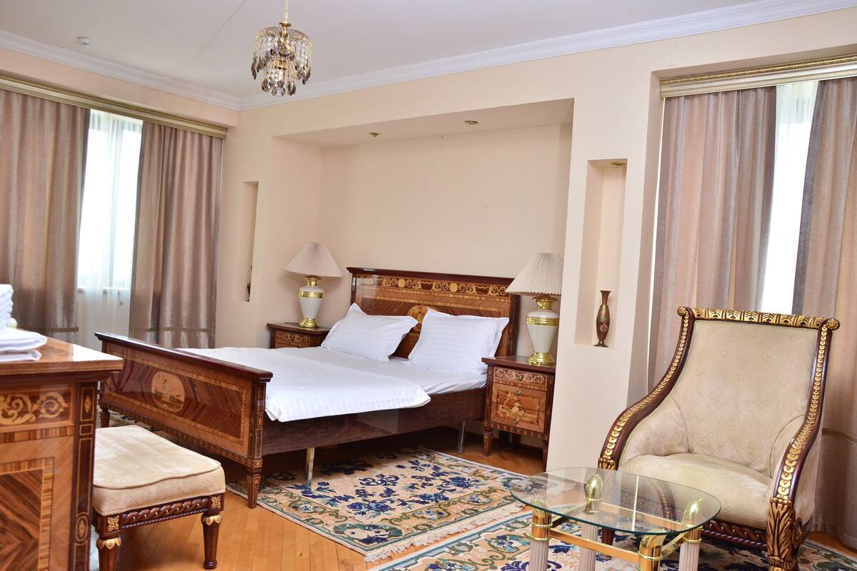 A hotel room with a large bed, a wooden nightstand with a lamp, a glass coffee table, a chair, and a decorative rug.
