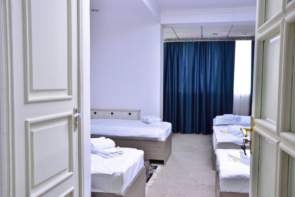 A hotel room with three single beds, a large window with blue curtains, and a carpeted floor.