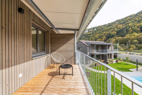 A wooden deck with a chair and table, a balcony with a railing, and a view of a mountain and a swimming pool. - Nadou – Lupus 5 Holiday Home in the Upper Danube Valley (Beuron)