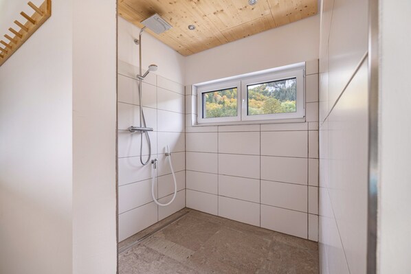 A small, tiled bathroom with a shower, a window with a view of trees, and a wooden ceiling.