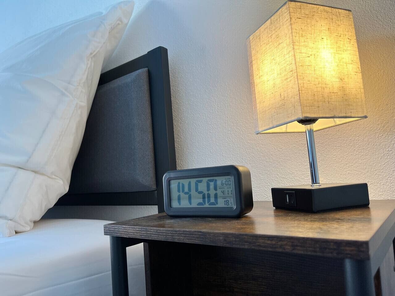 Une horloge numérique sur une table de chevet affiche 14 h 50, une lampe et un lit avec un oreiller et une tête de lit.